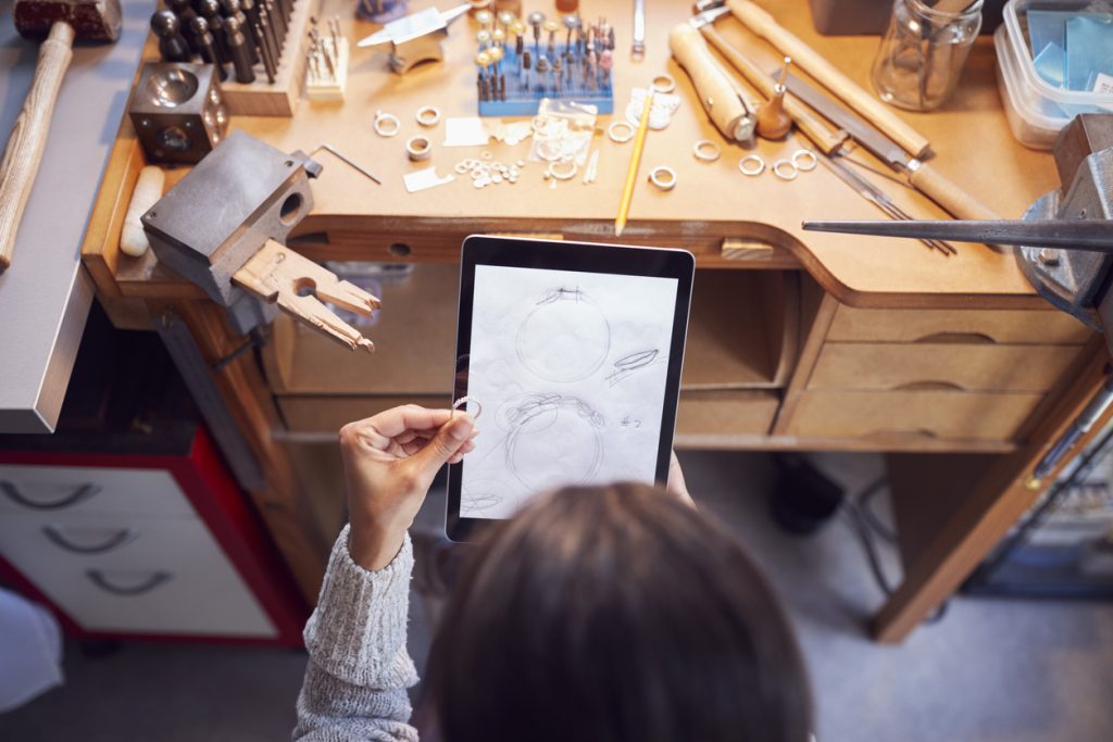 Overhead View Of Female Jeweller Comparing Ring With Drawn Design On Digital Tablet In Studio.